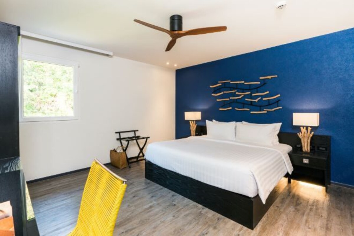 Un dormitorio moderno con una cama grande, un ventilador de techo, una lámpara fijada a la pared y una ventana con vista a la vegetación.