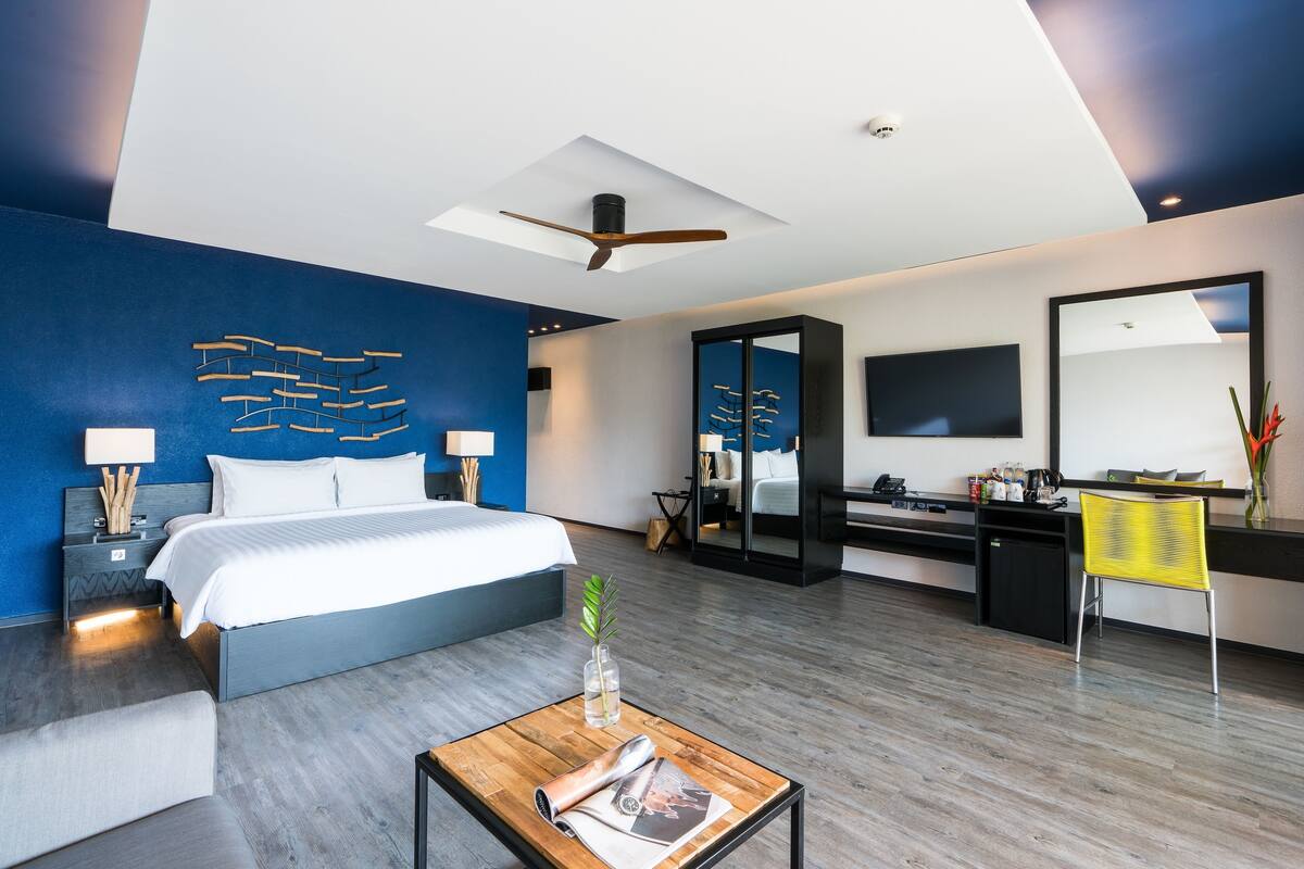 A modern hotel room with a large bed, a desk, a mirror, and a ceiling fan.