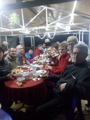 Un grupo de personas sentadas alrededor de una mesa redonda con diversos platos y utensilios, bajo una estructura parecida a una carpa con armazón metálico y luces colgantes.