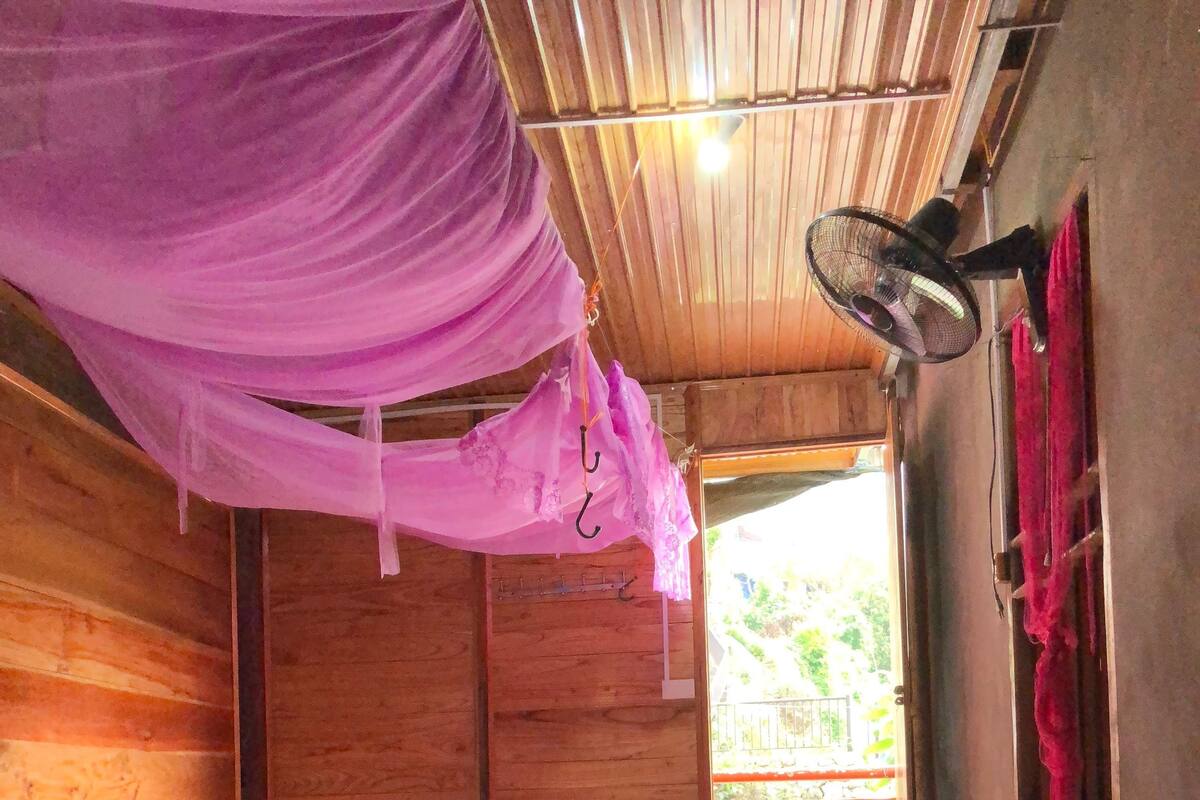 A wooden room with a bed, a hanging mosquito net, and a fan.