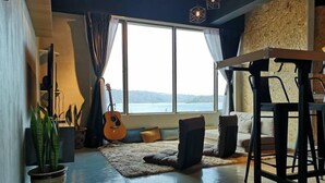A cozy living room with a large window, a guitar, a sofa, and a bar area. - Homy Seafront Hostel (Kota Kinabalu)