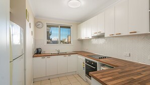 Een compacte keuken met witte kastjes, een houten werkblad, een inbouwoven en een raam met zonwering.