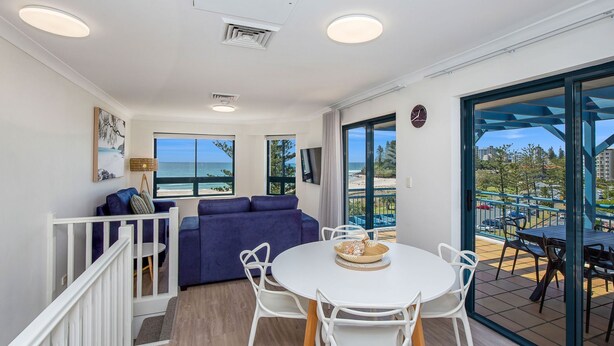 Una habitación con balcón, una mesa blanca con sillas, un sofá azul y vistas a la playa.