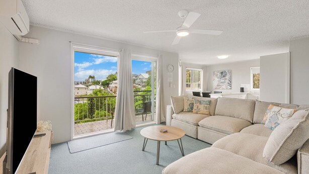 Un salon moderne avec un canapé, une table basse et un téléviseur. Il y a un balcon qui offre une vue sur le quartier.