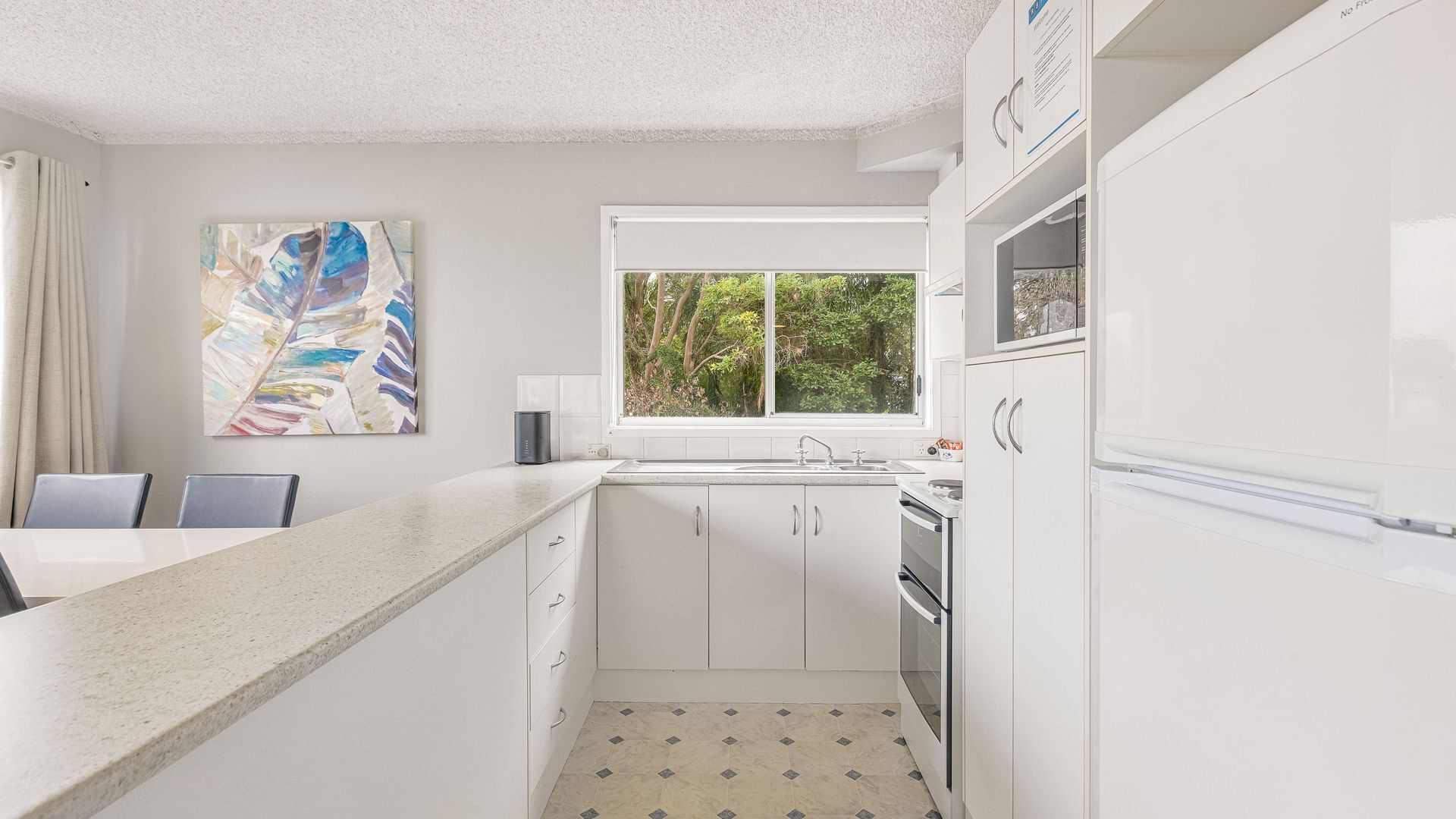 A modern kitchen with white cabinetry, a granite countertop, a window with a view of greenery, and a painting on the wall.