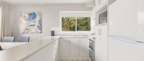 A modern kitchen with white cabinetry, a granite countertop, a window with a view of greenery, and a painting on the wall.