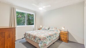 Une chambre à coucher avec un lit, une table de chevet, une commode en bois et une fenêtre avec des rideaux.