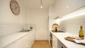 A modern kitchen with white cabinetry, a wooden floor, a wall clock, a sink, a cutting board, and a bottle of wine.