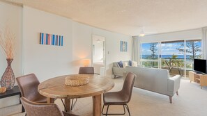 Ein modernes Wohnzimmer mit einem runden Holztisch, zwei Ledersesseln, einem Sofa und einem Fernseher. An den Wänden befinden sich abstrakte Gemälde und durch große Fenster ist der Blick auf den Ozean möglich.