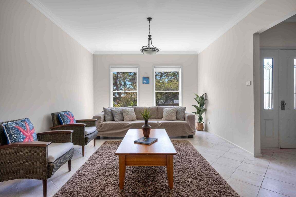 A spacious living room with a beige sofa, two armchairs, a wooden coffee table, and a large rug. There are two windows with white frames and a ceiling fan.