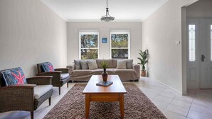 Un salon spacieux avec un canapé beige, deux fauteuils, une table basse en bois et un grand tapis. Il y a deux fenêtres avec des cadres blancs et un ventilateur de plafond.
