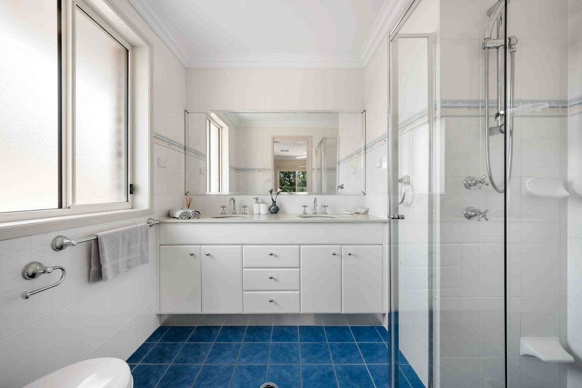 A modern bathroom with a large mirror, white cabinetry, a blue-tiled floor, and a glass-enclosed shower area.
