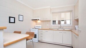 A compact kitchen with white cabinets, a stove, a sink, and a window. There are two wicker chairs and a small table.