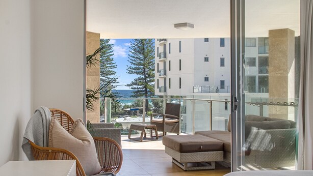 A balcony with wicker furniture, a cushioned bench, and a view of a building and ocean.