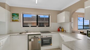 Una cocina moderna con armarios blancos, horno integrado, lavavajillas de acero inoxidable y ventana con vistas a una casa y árboles.
