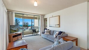 A modern living room with a grey sofa, a wooden coffee table, and a flat-screen TV. A large sliding glass door leads to an outdoor area with a view of the ocean and parked cars. - Kooringal Unit 3 Right on Greenmount Beach Coolangatta with Wi-Fi Included (Coolangatta)