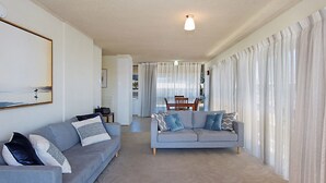 A living room with a grey sofa, a dining table with chairs, and a kitchen area in the background. - Kooringal Unit 3 Right on Greenmount Beach Coolangatta with Wi-Fi Included (Coolangatta)