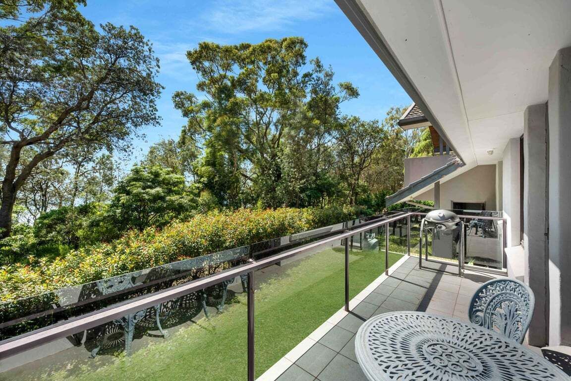 Un balcón con barandilla de vidrio, mobiliario exterior y vista a un jardín con árboles y arbustos.