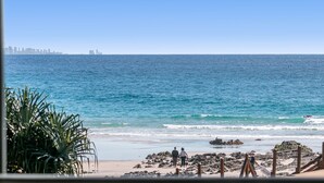 Une plage au rivage rocheux, avec la silhouette d’une ville en arrière-plan et un ciel bleu dégagé.