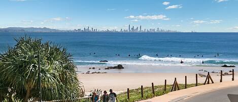 A coastal road with a beach and ocean view, a city skyline in the distance, and a palm tree on the left.