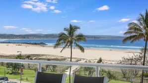 Une vue sur la plage depuis un balcon doté d’une balustrade, avec des palmiers et un ciel dégagé.