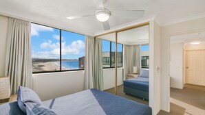 Un dormitorio con cama, vista a la playa, ventilador de techo y puerta que da a otra habitación.