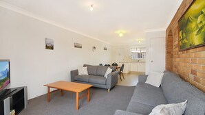 A compact living space with a grey sofa, a wooden coffee table, and a TV on a stand. A kitchen is visible in the background. - Allamanda Court Unit 6 neat and tidy unit handy to Tweed Heads Bowls Club (Tweed Heads)