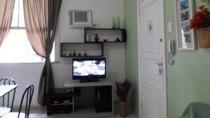A living room with a television on a stand, a small shelf with decorative items, a door, and a window with curtains.