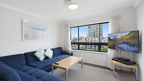 A modern living room with a blue sofa, a small wooden table, a flat-screen TV, and a large window offering a view of a high-rise building. - Calypso Plaza Resort Unit 417 Two bedroom unit in resort style complex central Coolangatta (Coolangatta)