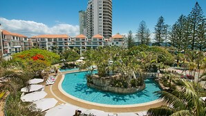 Et resort med en central pool omgivet af palmer og liggestole, en høj bygning i baggrunden og en klar blå himmel.