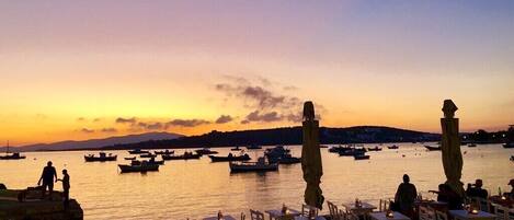 A waterfront restaurant with outdoor seating overlooking the sea, boats in the background, and a sunset sky.