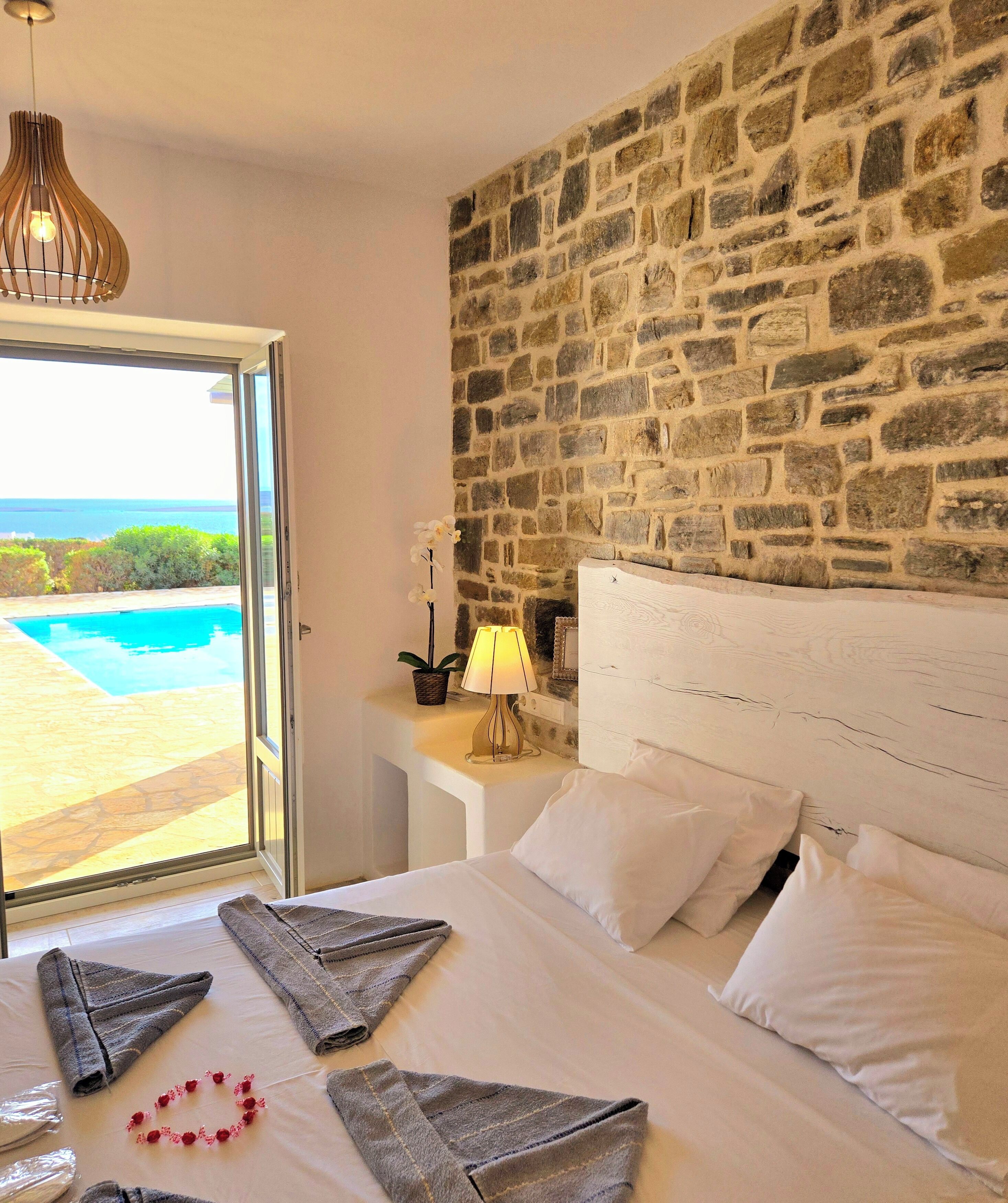 Een hotelkamer met een stenen muur, een bed met witte lakens, uitzicht op zee, en een hanglamp.