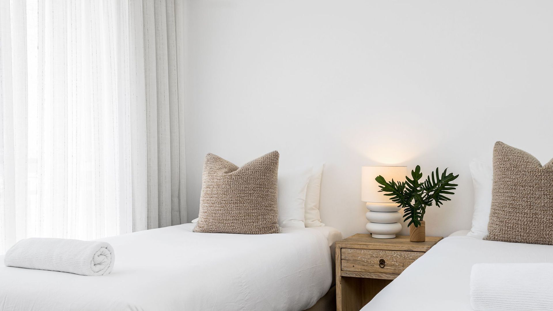 A hotel room with two beds, a nightstand with a lamp and a plant, and a window with curtains.