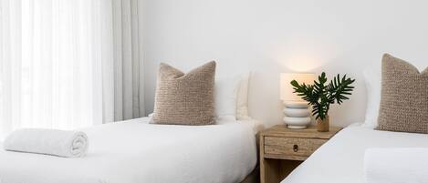 A hotel room with two beds, a nightstand with a lamp and a plant, and a window with curtains.