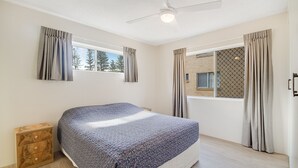 Une chambre à coucher avec un lit, une table de chevet en bois, un ventilateur de plafond et une fenêtre avec des rideaux.