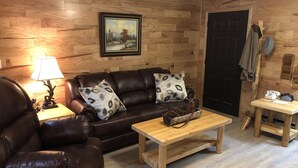 A cozy living room with a brown leather sofa, two patterned pillows, a wooden coffee table with pinecones, a framed landscape painting, and a wooden door with a coat rack. - HOT TUB COTTAGE near Cave Run Lake, Kentucky, Red River Gorge,Morehead Kentucky (Wellington)