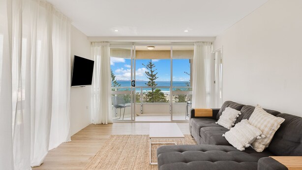 A modern living room with a grey sofa, white cushions, a flat-screen TV, and a view of the ocean through large glass doors.