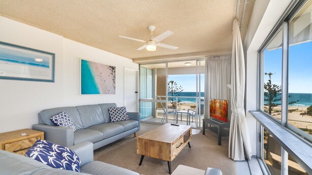A modern living room with a grey sofa, coffee table, and artwork on the walls. There is a balcony with a view of the beach. - Kooringal Unit 20 Located right on Greenmount beach with ocean views (Coolangatta)