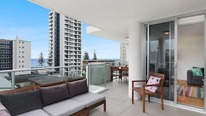 Un balcon avec un canapé et des fauteuils, une porte vitrée donnant sur une pièce, et une vue sur un immeuble imposant et des arbres.