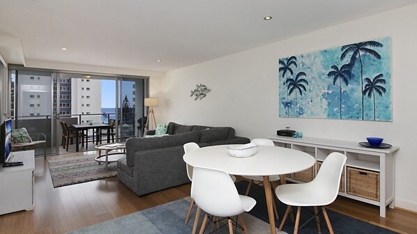 En moderne stue med en grå sofa, et hvidt spisebord og stole. Der er en balkon med udsigt til bygninger og træer.