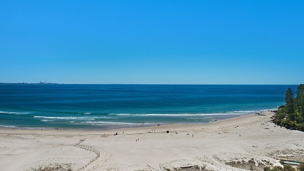 A wide sandy beach with gentle waves, a clear blue sky, and a distant city skyline. - Reflections On The Sea Unit 1501 On Coolangatta Beach, Ocean and Coastline views with Wi-Fi Inc. (Coolangatta)