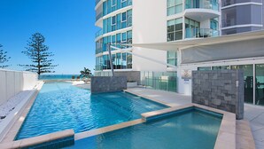 Un área de piscina moderna con agua azul cristalina, un edificio blanco con grandes ventanales y vista al océano.