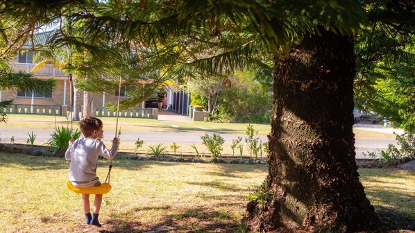 Un enfant sur une balançoire, sous un arbre dans un parc, avec un bâtiment en arrière-plan.