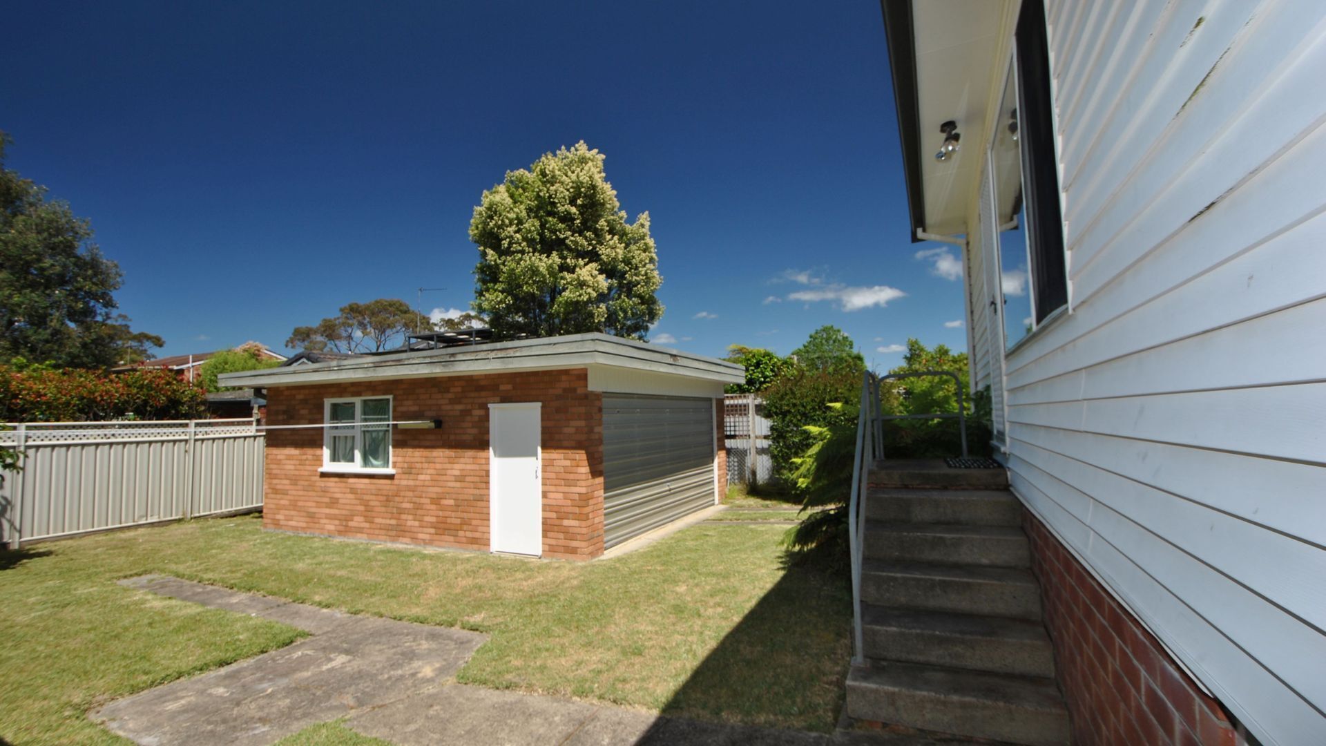 A small brick garage with a white door, a metal fence, and a tree in the background.