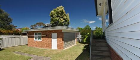 A small brick garage with a white door, a metal fence, and a tree in the background.