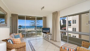 Een ruim balkon met een televisie, rieten meubels en uitzicht op het strand en gebouwen.