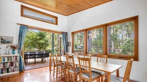 Un coin repas avec une table et des chaises en bois, de grandes fenêtres donnant sur la forêt et une bibliothèque remplie de livres.