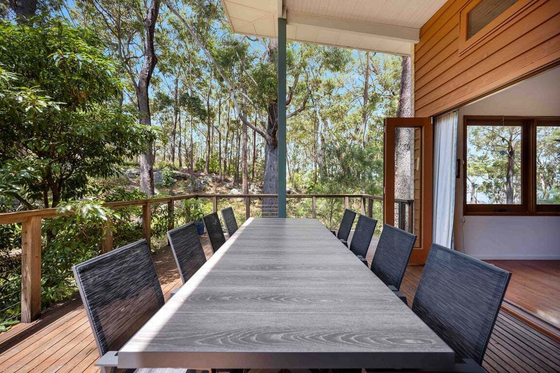 A long dining table with chairs, set on a wooden deck with a view of a forest.
