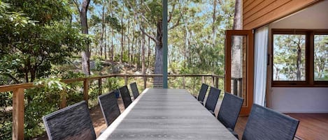 A long dining table with chairs, set on a wooden deck with a view of a forest.