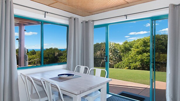 Un espace repas comprenant une table et des chaises, de grandes fenêtres avec des rideaux, et une vue sur la verdure et une statue.